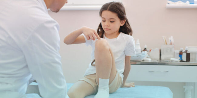 A young girl points to her swollen knee while sitting on an exam table, during a pediatric consultation—representing juvenile arthritis symptoms and early diagnosis.