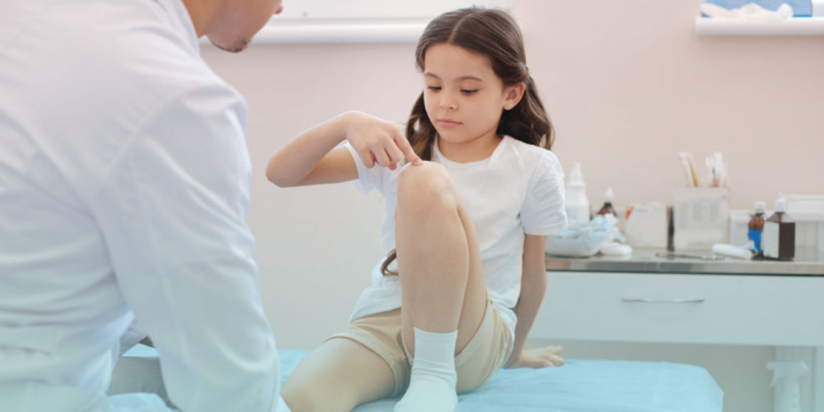 A young girl points to her swollen knee while sitting on an exam table, during a pediatric consultation—representing juvenile arthritis symptoms and early diagnosis.